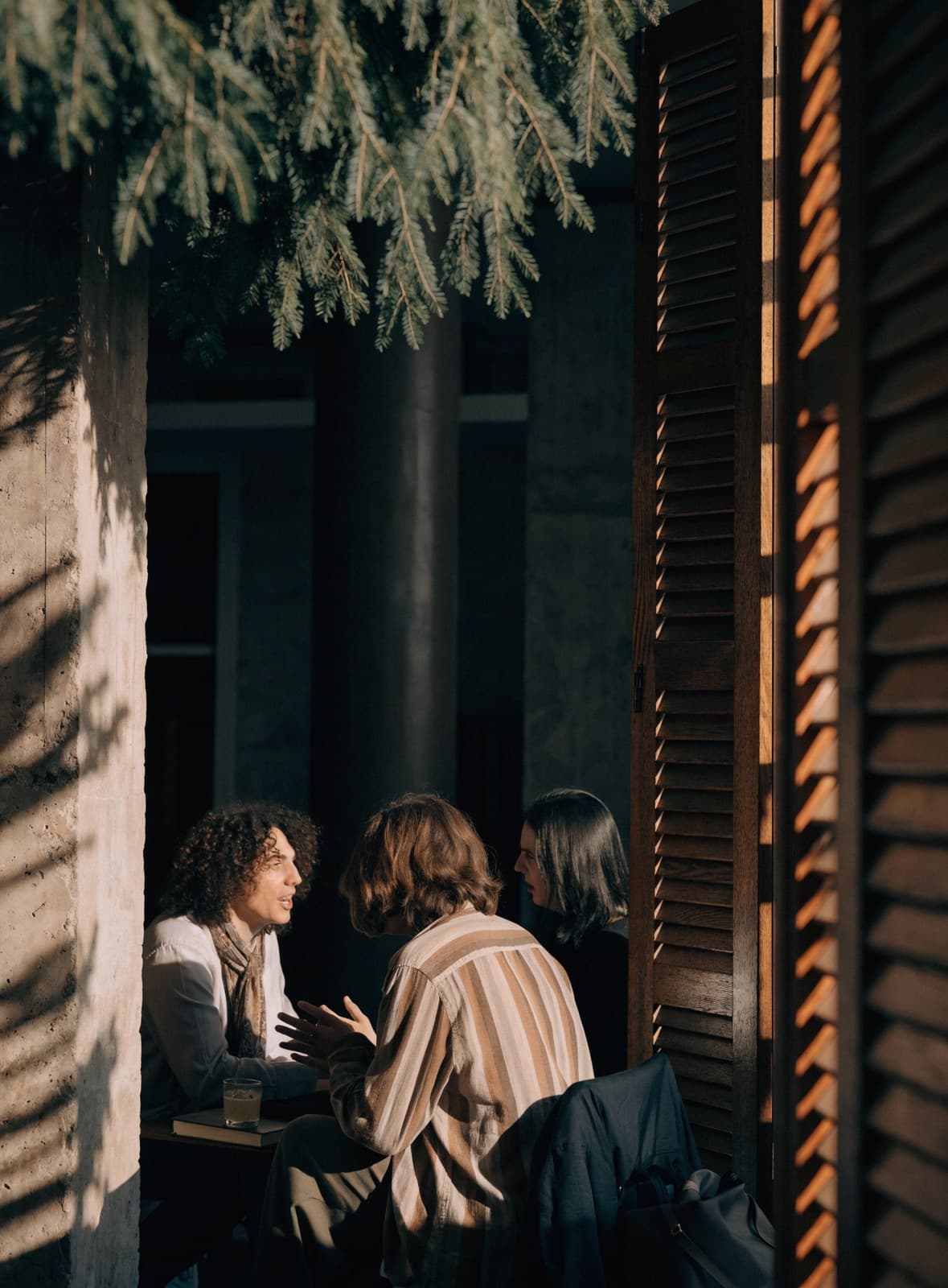 Three people seated close together in conversation, warm light coming through a wooden shutter.